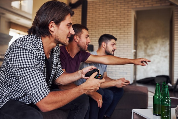 Side view. Group of friends have fun playing console game indoors at living room.