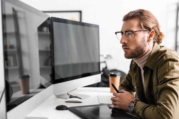 Photo side view of handsome 3d artist holding stylus near graphics tablet and looking at computer monitors