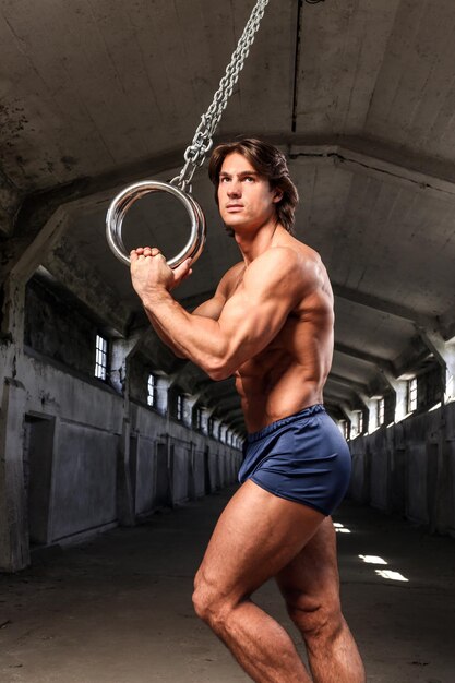 Side view of a handsome athlete with muscular body posing with gymnastics rings in the abandoned industrial building