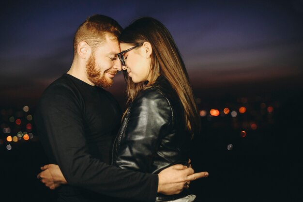 Side view of handsome bearded guy keeping eyes closed and showing fuck off gesture while standing on blurred background of night city and embracing lovely young woman