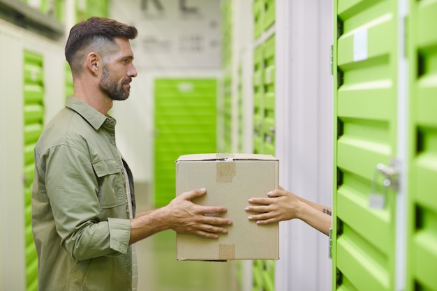 Photo side view at handsome bearded man holding cardboard box and handing in to woman while packing self storage unit, copy space