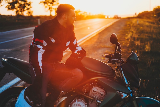 Vista laterale dell'uomo barbuto bello in equipaggiamento protettivo che si siede in piedi sul sedile della moto lungo la strada con il casco in mano su sfondo sfocato retroilluminato del percorso del paese