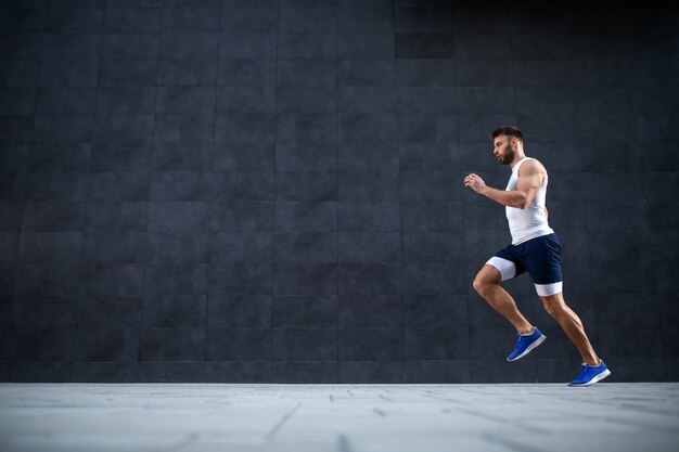 Vista laterale dell'uomo caucasico bello muscoloso in forma che corre veloce all'aperto. sullo sfondo è il muro grigio.