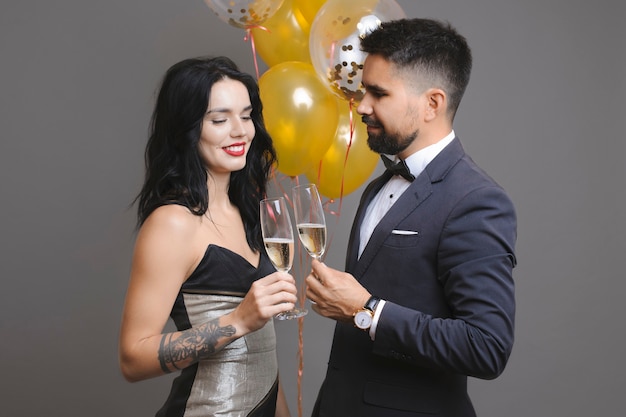 Side view of handsome man in suit and beautiful woman in evening dress smiling and clinking glasses of champagne while standing near bunch of balloons on gray background