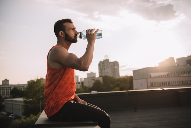 Вид сбоку на красивого спортсмена, пьющего воду из спортивной бутылки на крыше