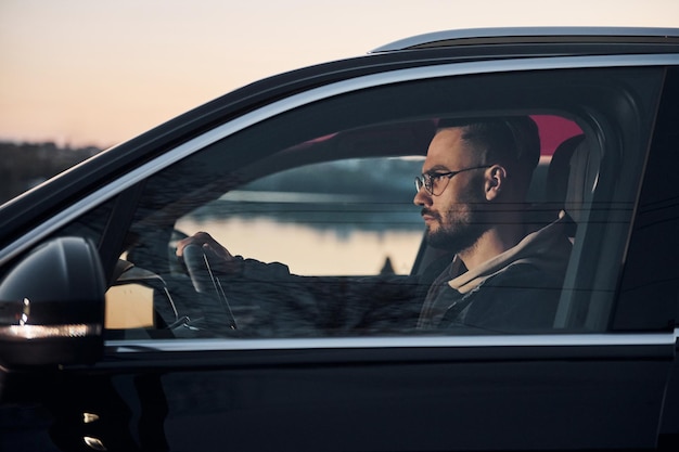 Photo side view of handsome unshaved man in fashionable clothes that riding his automobile