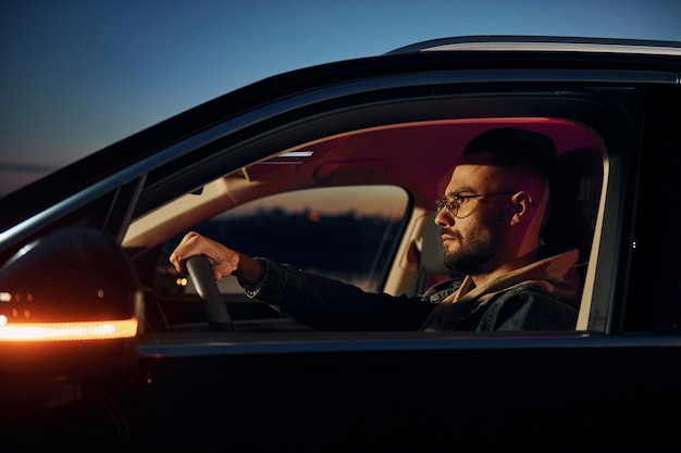 Side view of handsome unshaved man in fashionable clothes that riding his automobile