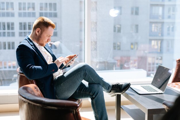 Side view of handsome young businessman wearing fashion suit is using mobile phone in modern office room at the wooden desk on background of large window laptop on table Concept of office working