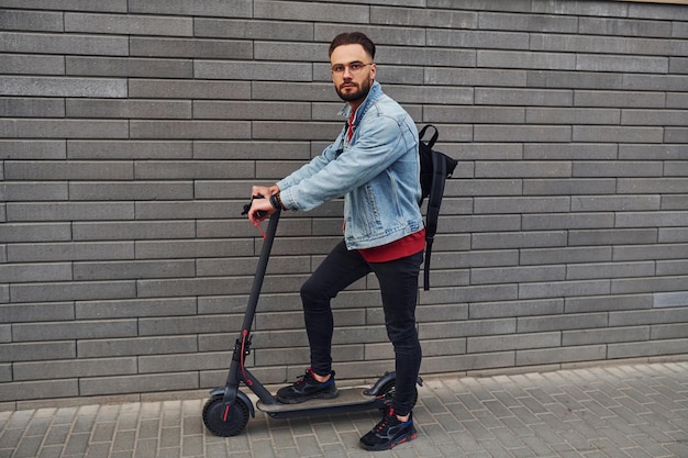 Side view of handsome young guy in casual clothes that riding electric schooter outdoors at sunny daytime