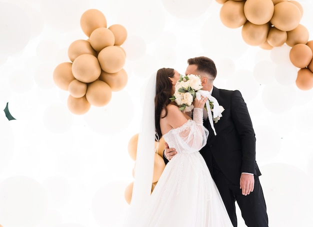 Вид сбоку счастливой пары в свадебной одежде, целующейся и закрывающей лицо букетом цветов, стоя на фоне свадебной арки, украшенной гелиевыми шарами