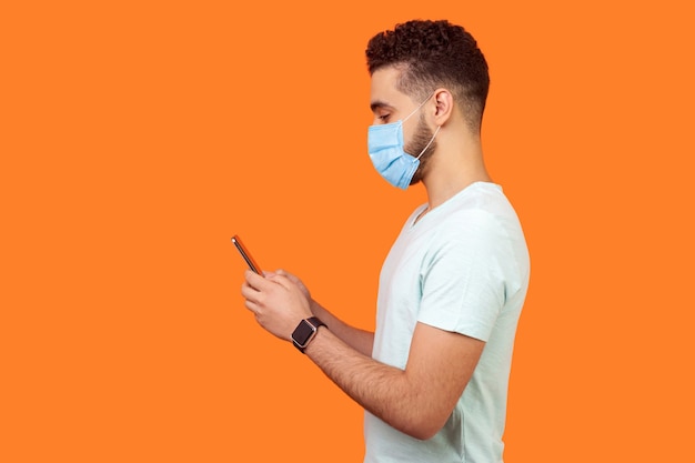 Vista laterale dell'uomo bruna felice con maschera medica chirurgica che sorride mentre usa il cellulare, chatta nei social media, godendo del servizio mobile. copia spazio vuoto per pubblicità, studio indoor