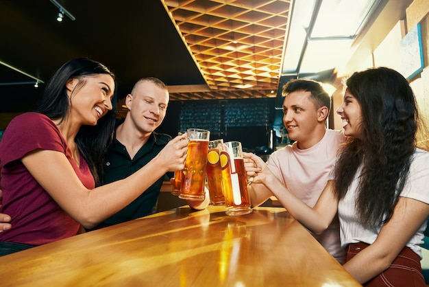 Side view of happy company sitting together in pub and drinking beer at weekend. Cheerful young male and female looking at each other, talking and laughing in bar. Concept of fun and happiness.