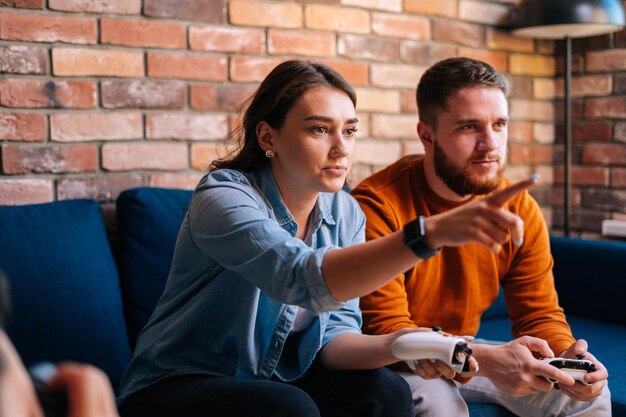 Side view of happy focused young couple holding controllers and playing video games on console sitting together on couch at cozy living room. Concept of leisure activity of lovers at home.