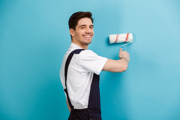 Vista laterale della parete della pittura del costruttore maschio felice con il rullo della vernice mentre sopra la parete blu
