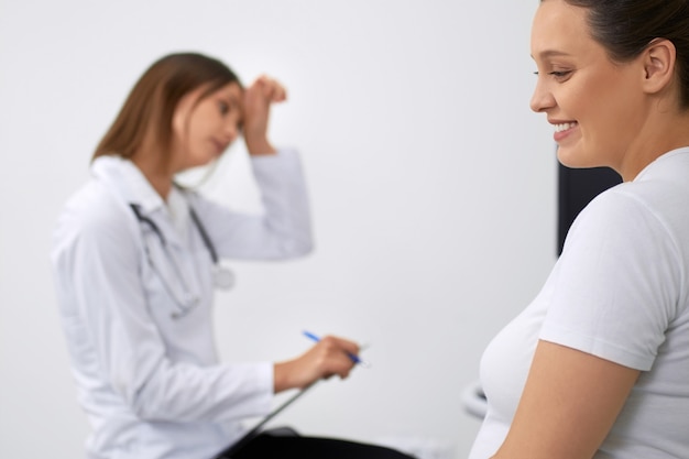 Side view of happy pregnant woman sitting at medical center while gynecologist providing professional diagnostic