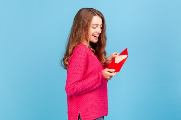 분홍색 스웨터를 입고 편지나 연하장을 읽고 봉투를 들고 웃고 즐거운 소식을 기뻐하는 행복한 여성의 측면. 파란색 배경에 고립 된 실내 스튜디오 촬영입니다.