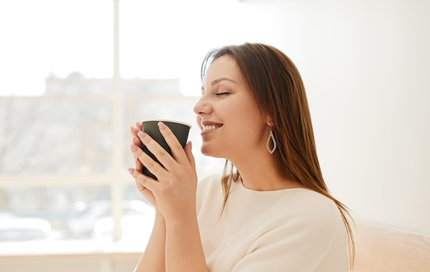 家で朝を楽しみながら広く笑っている熱い芳香のコーヒーのマグカップと幸せな若い女性の側面図