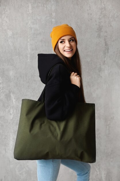 A side view of a hipster model girl in a black sweatshirt and yellow hat with a shopper bag.