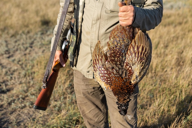 Photo side view of hunter carrying dead pheasant while walking on field