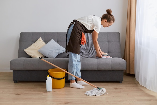 Photo side view image of young woman cleaning her home wearing jeans and brown apron housewife doing her household chores at home in light living room near window