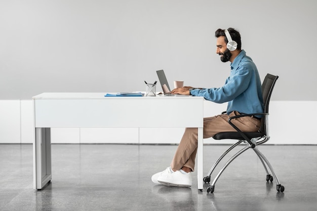 Side view of indian businessman in headphones working on laptop making video call sitting at desk