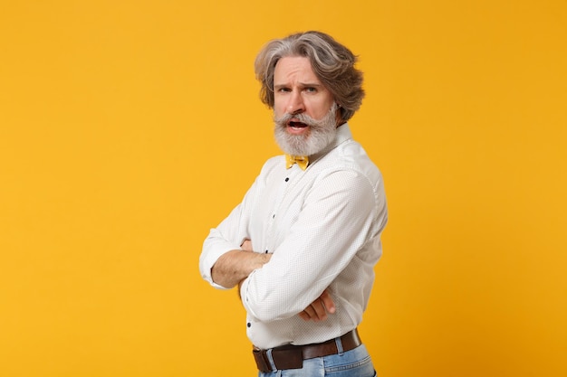 Side view of irritated elderly gray-haired mustache bearded man\
in white shirt bow tie posing isolated on yellow orange background.\
people lifestyle concept. mock up copy space. holding hands\
crossed.