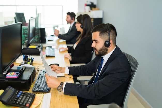 Side view of a latin man with a headset reading instructions and holding some paperwork. Sales representative working on telemarketing and customer service