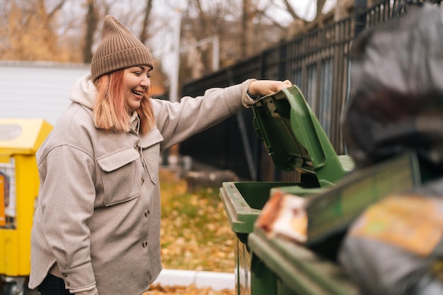 Side view of laughing overweight female wearing autumn hat and jacket opening trash can and looking inside. Attractive young homeless female looking for food in street garbage can in cloudy day.