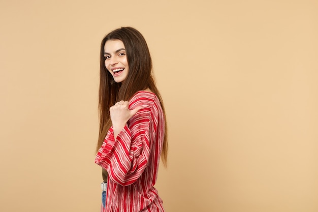 Vista laterale della giovane donna ridente in abiti casual che guarda la telecamera, puntando il pollice da parte isolato su sfondo beige pastello in studio persone sincere emozioni, concetto di stile di vita. mock up copia spazio.