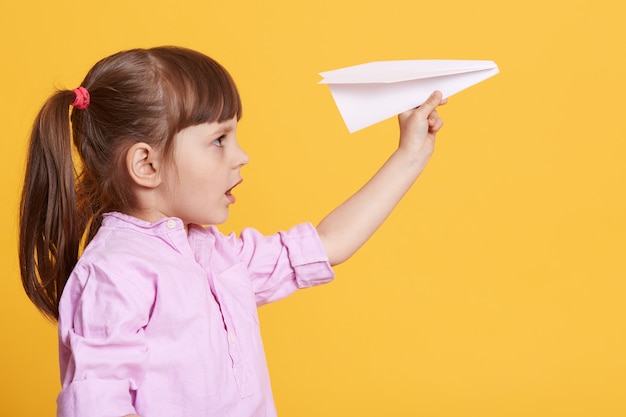 Side view of little cute female kid posing with white paper plane in hands