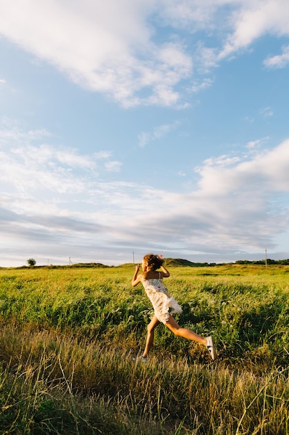 Side view of little happy girl in dress running with kite in green field