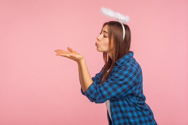 Side view of lovely pretty girl with angelic saint nimbus blowing sensual air kiss over palms flirting and expressing love amorous romantic feelings indoor studio shot isolated on pink background