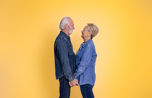 Side view of loving senior man and woman holding hands and romantically looking at each other while standing isolated on yellow background