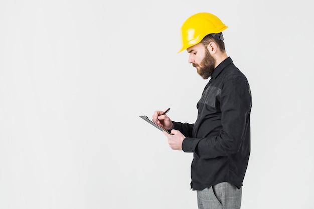 Photo side view of male architect wearing yellow hardhat writing on clipboard