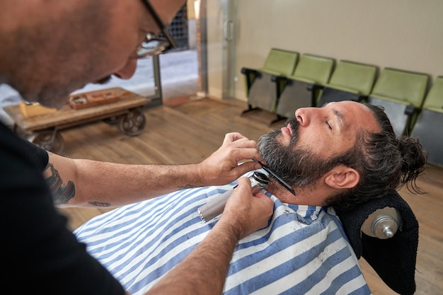 Photo side view of male barber cutting beard of client with trimmer while working in modern hairdressing salon