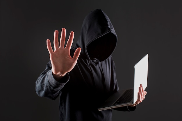 Side view of male hacker holding laptop and putting hand up as stop