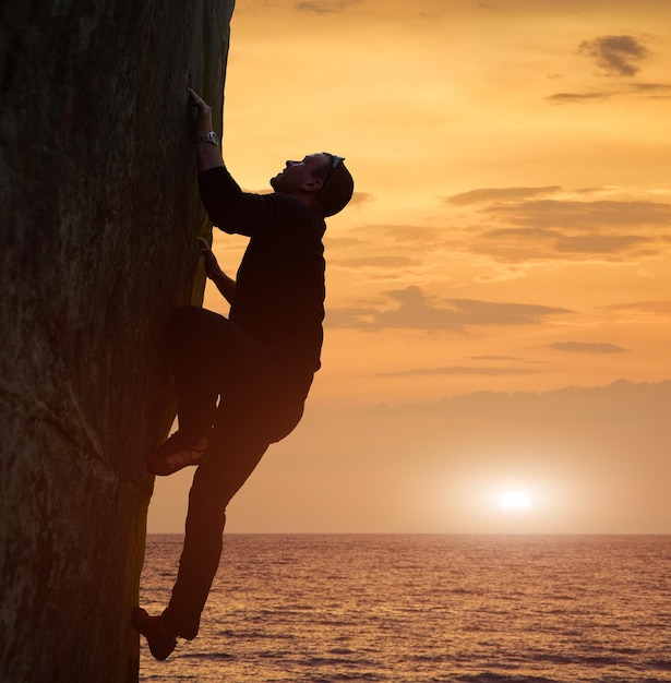 Side view of man in black climbing on rock without safety harness over sea in twilight Recreational activity in nature