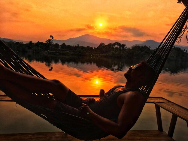 Photo side view of man lying on hammock against lake during sunset