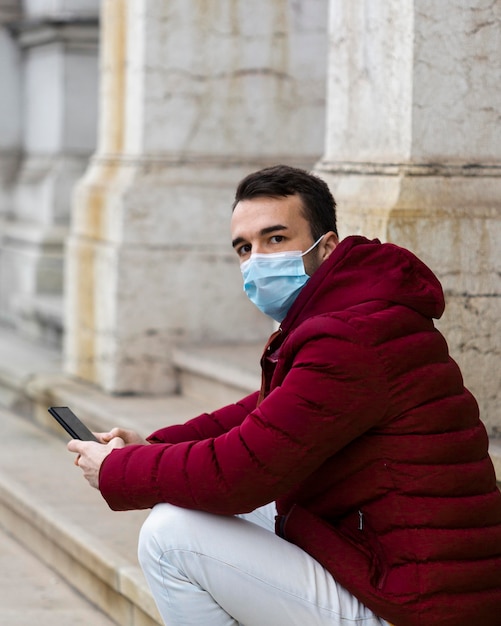 Photo side view of man sitting outdoors with medical mask and holding smartphone