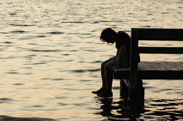 Photo side view of man sitting on pier over lake during sunset