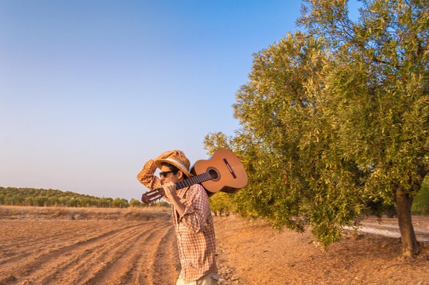 Foto vista laterale di un uomo con una chitarra acustica che cammina tra gli alberi sul campo