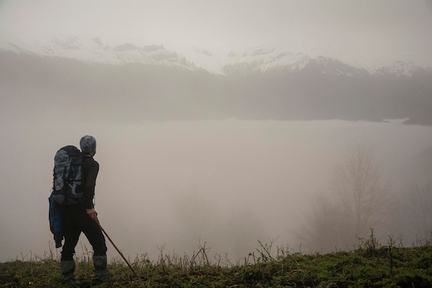 Foto vista laterale di un uomo con uno zaino che cammina sul campo vicino al lago durante il tempo nebbioso