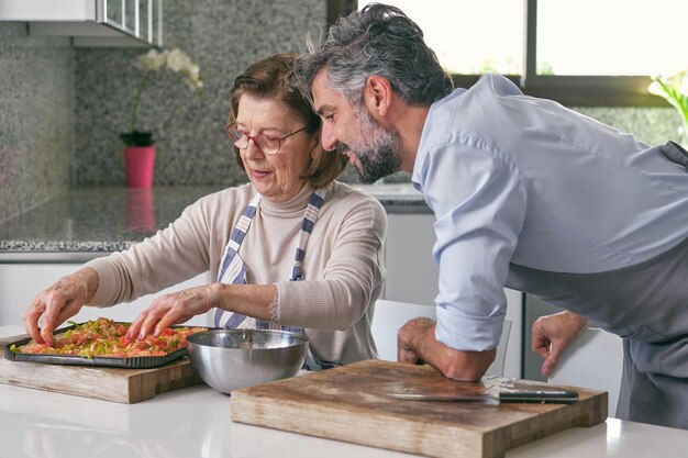 회색 머리카락을 가진 성숙한 수염을 가진 히스패닉 남성과 집에서 맛있는 이탈리아 음식을 준비하는 <unk>을 입은 노인 여성의  ⁇ 보기