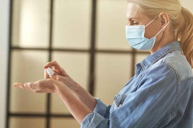 Foto vista laterale di una donna bionda matura con una maschera protettiva che si pulisce le mani usando un antibatterico