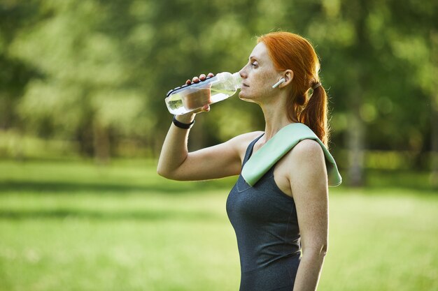 Vista laterale della donna matura di forma fisica della testarossa con l'asciugamano sull'acqua potabile della spalla nel parco dopo l'esercizio