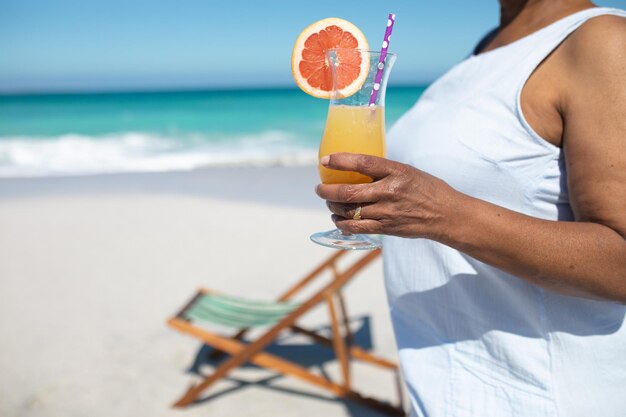 Foto vista laterale a metà sezione di una donna afroamericana anziana su una spiaggia in piedi al sole, con un cocktail in mano, con una sdraio sulla spiaggia, cielo blu e mare calmo sullo sfondo
