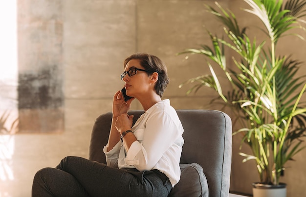 Side view of a middleaged woman in formal wear sitting indoors and talking on a mobile phone