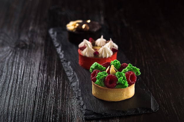 Side view on mini party tartlets with fresh fruit, chocolate, nuts and cream on black slate background. Sweet cakes