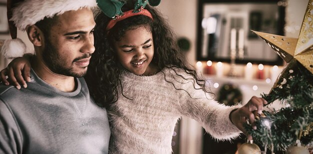 Side view of a mixed race man with his young daughter in their sitting room at Christmas, wearing festive hats, the man holding his daughter touching a Christmas tree