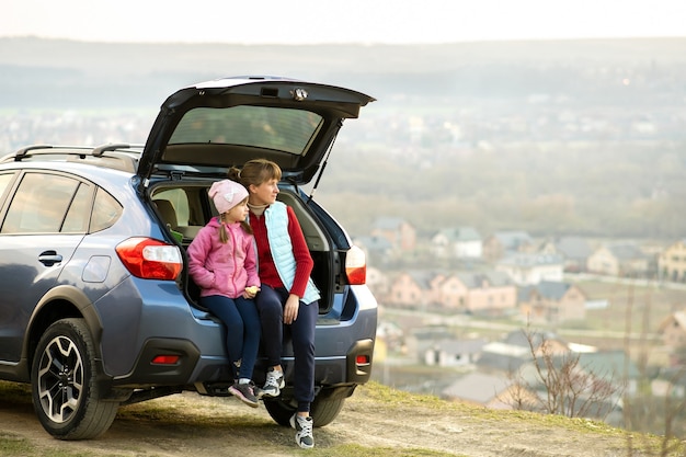 Side view of mother with daughter sitting in car trunk and looking on nature. Concept of resting with family on fresh air.